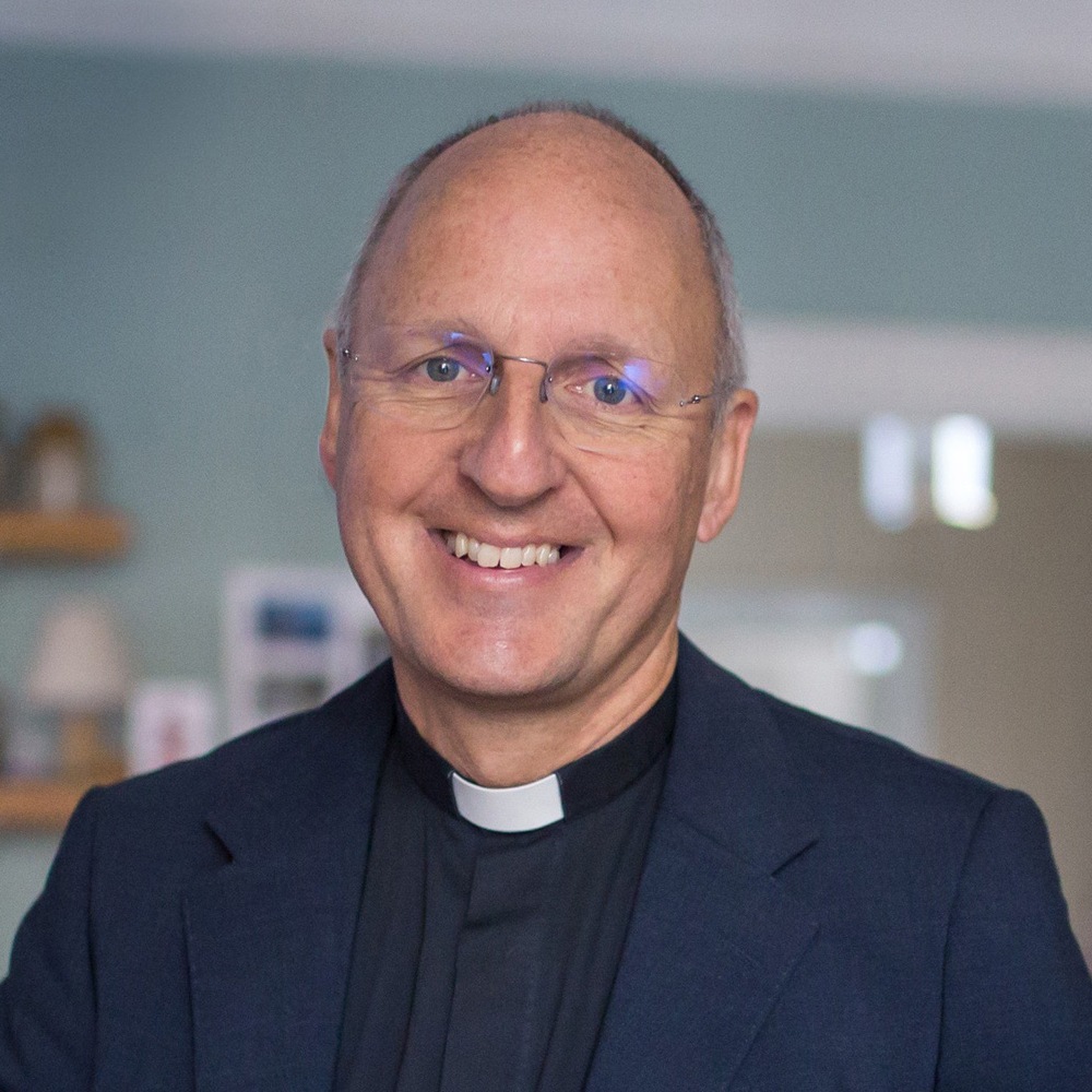 Head an shoulders portrait of a smiling man, wearing frameless glasses, and a black jacket/shirt and a clerical collar