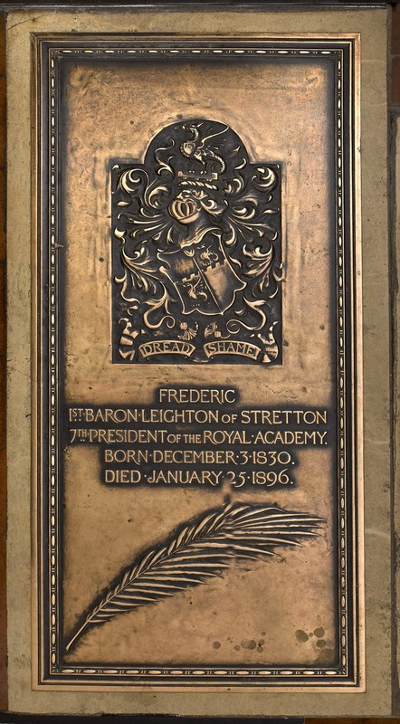 Brass panel with decorative frame; a heraldric family motto with dragon, helmet and shield, above the inscription, and a single feather