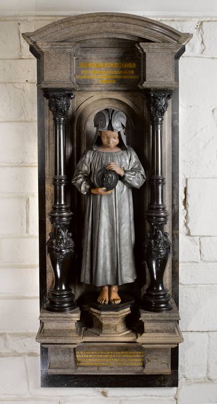 A barefoot child wearing a smock and helmet looks down at a meadllion he/she is holding with both handd - a portrait head of Randolph Caldecott. Either side, are black, ornate pillars within a stone surround, whcih carries an inscription in gold, top and bottom.