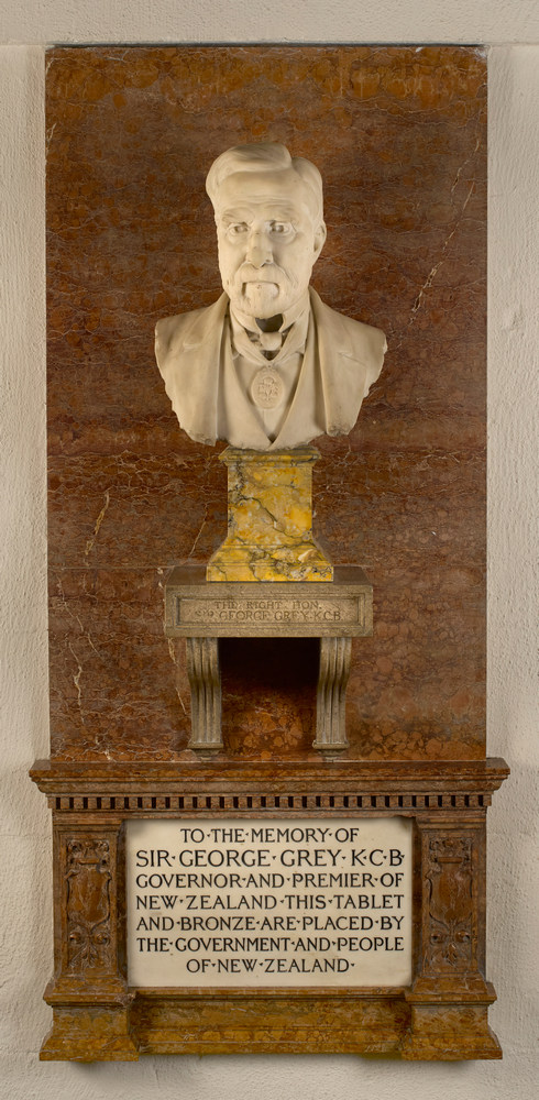 Monument showing a white marble portrait bust of a bearded man set on a yellow marble plinth, above a white marble inscription with black lettering - all against a dark-brick-red marble background