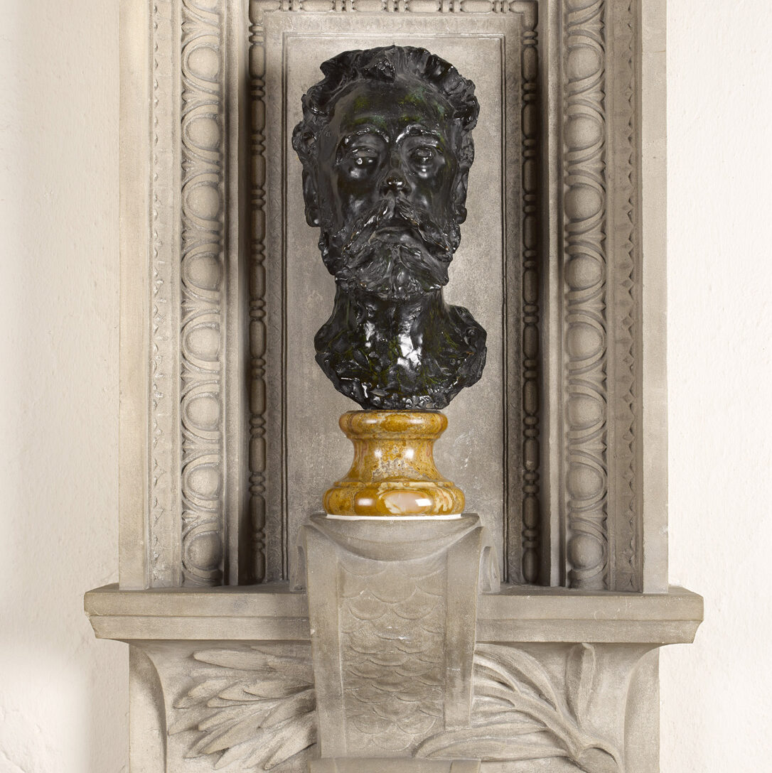 Bronze bust of bearded man on a yellow stone plinth, set in a decorative square-framed alcove .