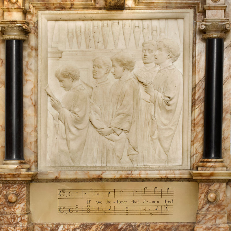 Relief panel in white marble showing five choristers in profile, singing from music held in their hands; organ pipes in the background. Under the panel , two bass staves of music with the words of Goss' anthem, 'If we believe that Jesus died'