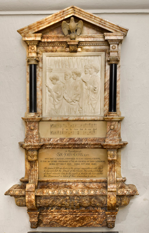 A marble wall monument wutha white marble relief panel showing choristirs singing, set above a stave of music: all framed by yellow marble with an inscription