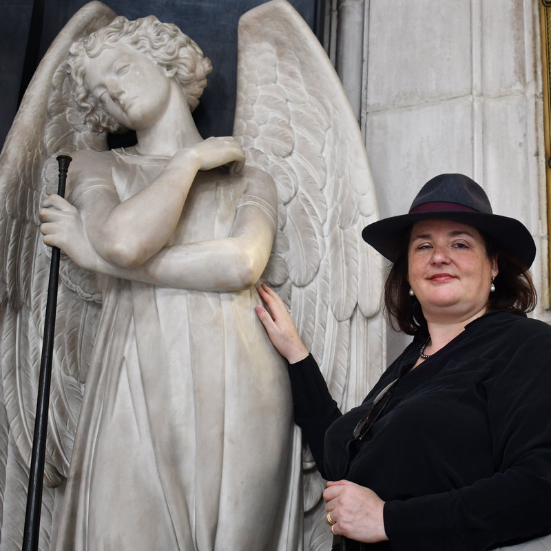 Woman dressed in black standing next to a tall angel sculpted in marble