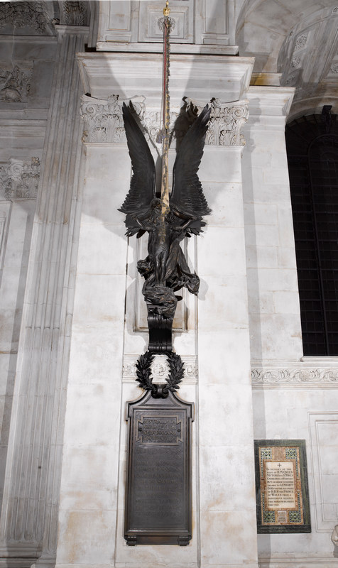 Tall wall monument showing bronze Christ crucified being borne by an angel wrapped in drapery and with huge wings