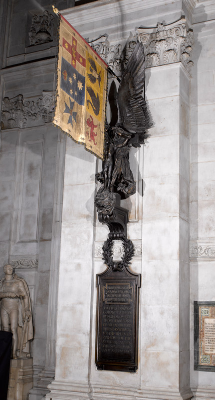 Side view of long wall monument showing bronze Christ crucified being borne by an angel wrapped in drapery and with huge wings, surmounted by a military standard, which hangs down in front.