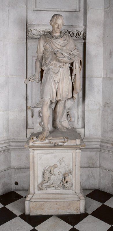 free-standing marble figure wearing a toga-like garment,holding a scroll in one hand and a keyn in the other, standing on a plinth with a prison scene sculpted in relief