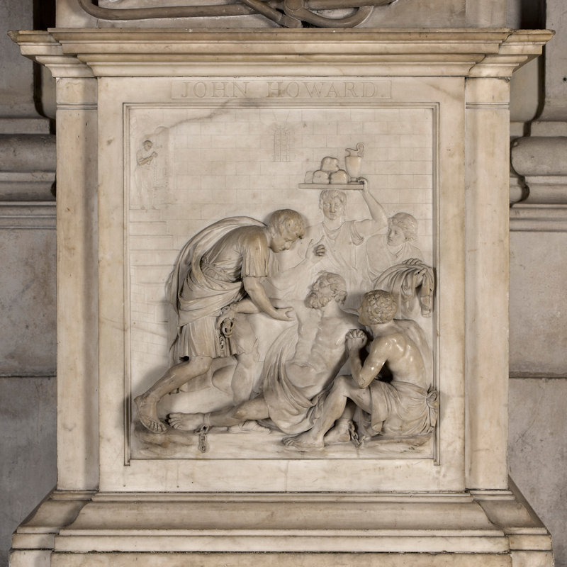 Sculpted relief panel showing a prisoner being helped by cvarius people, including someine with a tray of bread and wine on their head, while a jailer holding a large key looks on, top left corner.