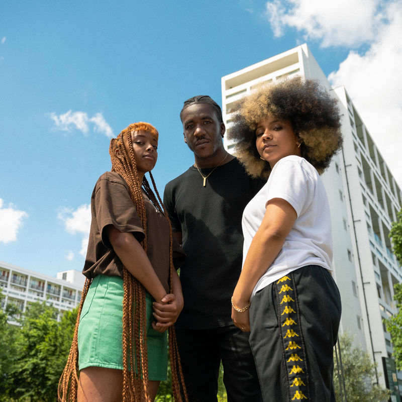 Three people photgraphed from low down standing in front of a block of flats, against a blue sky