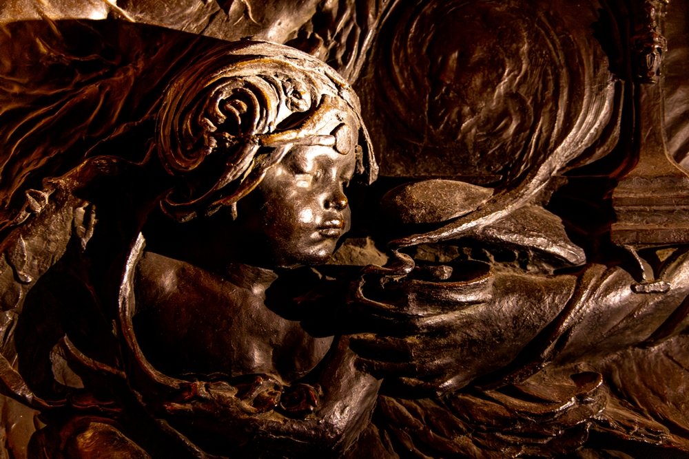 Close-up of bronze cherub, blowing smoke from a lamp or incense burner, in a rich brown/coppery glow.