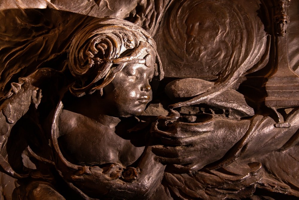 Close-up of bronze cherub, blowing smoke from a lamp or incense burner, in a brown/coppery glow.