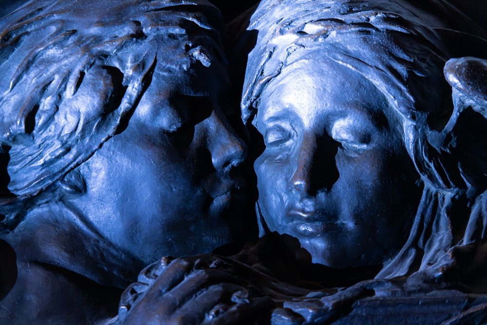 Close-up of two close up of two bronze heads , one whispering in the other's ear, in a blueish light